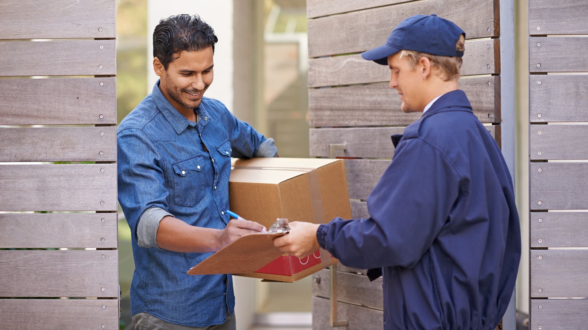 Delivery person hands a cardboard box to a person, both smiling, at a wooden doorway. Delivery person hands a cardboard box to a person, both smiling, at a wooden doorway.