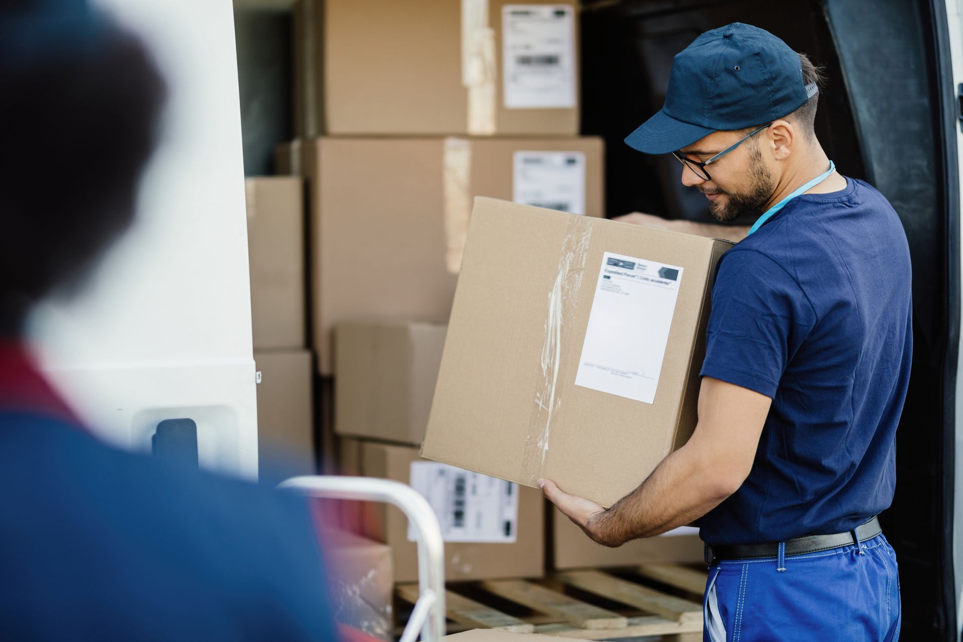 Delivery person loading boxes into a van. Wearing blue uniform, cap, and glasses.