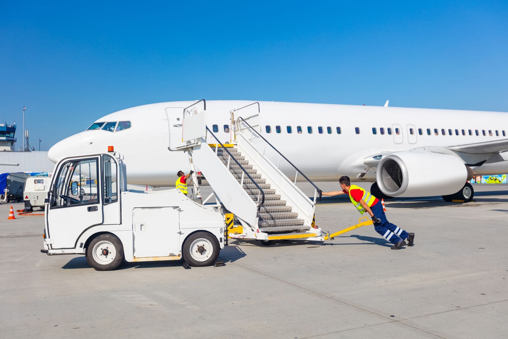 Two airport workers push a mobile staircase towards a white airplane on a sunny day.