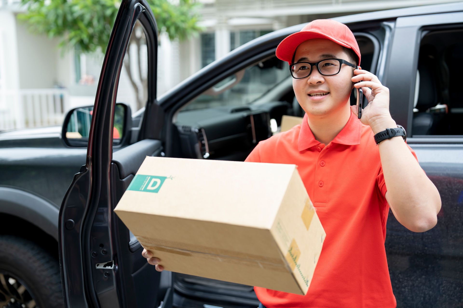 An Asian delivery man in a red uniform holding a cardboard package while talking on a mobile phone