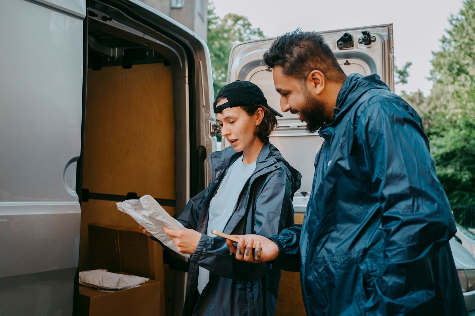 Two people in rain jackets review papers next to a van full of boxes, outdoors.