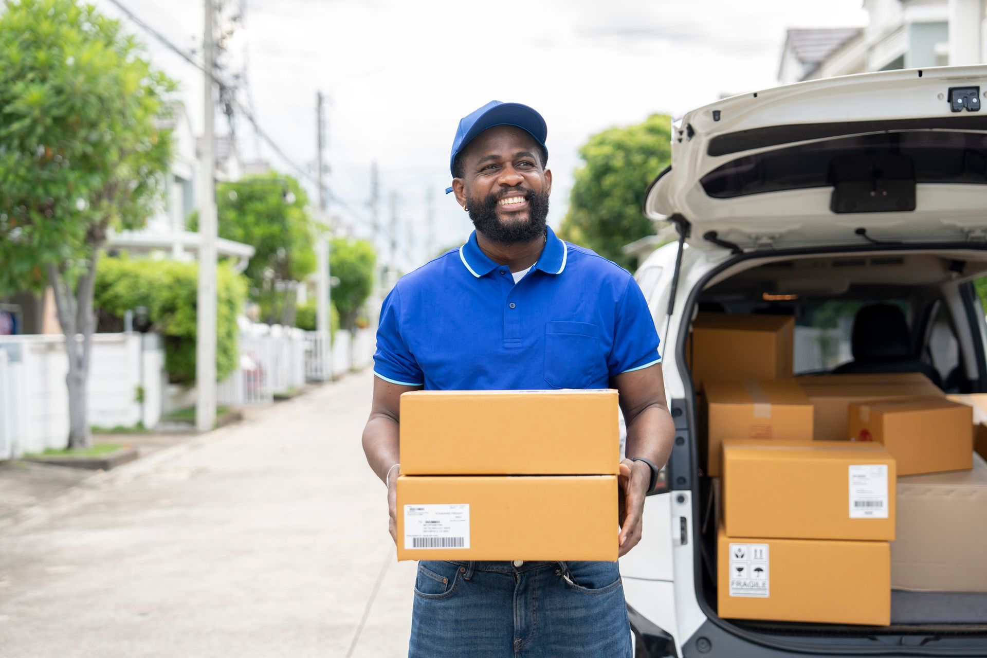 Delivery person holding packages near van, smiling. Blue shirt, cap. Suburban street.