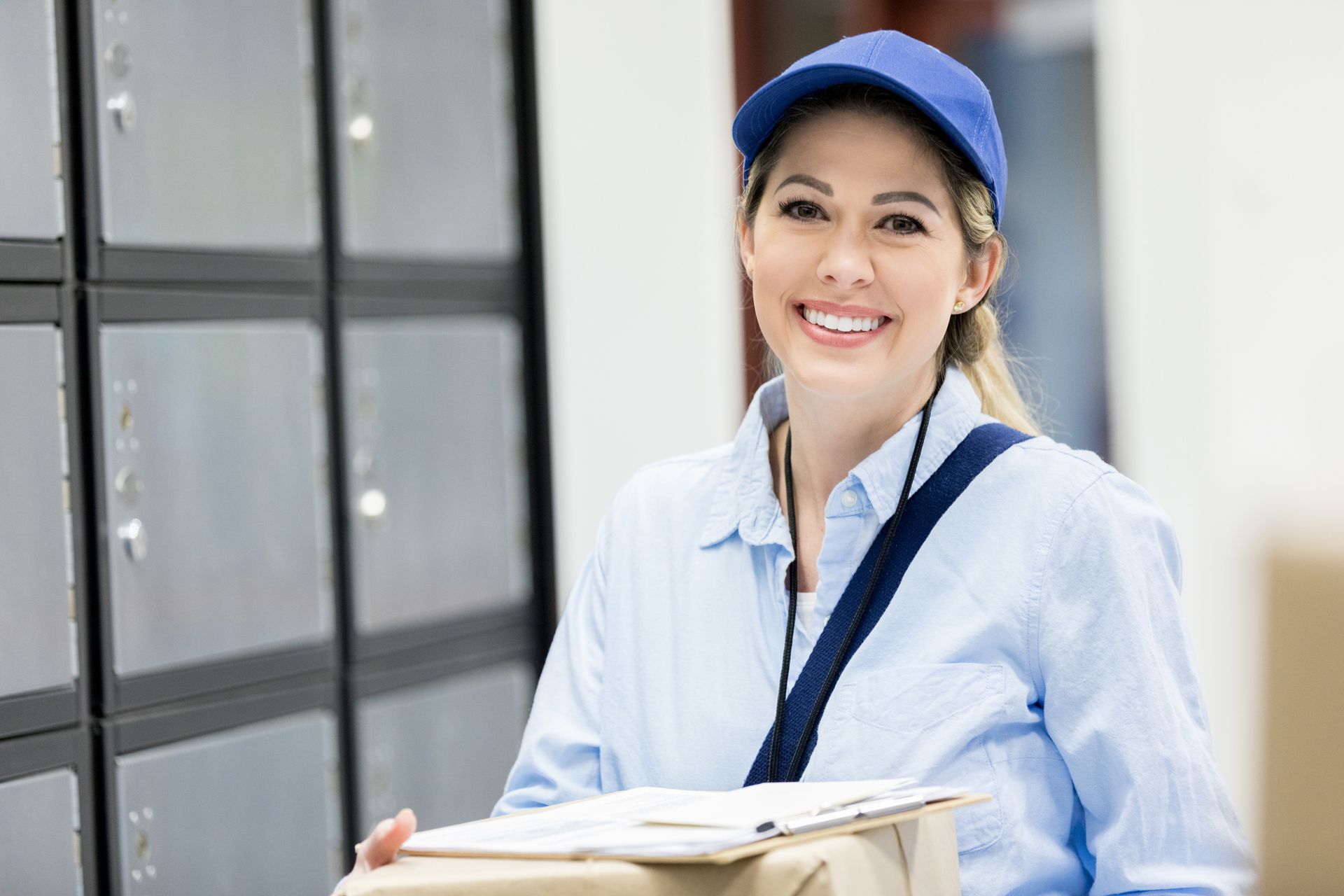 Smiling mail carrier in blue uniform, holding package, standing by mailboxes.
