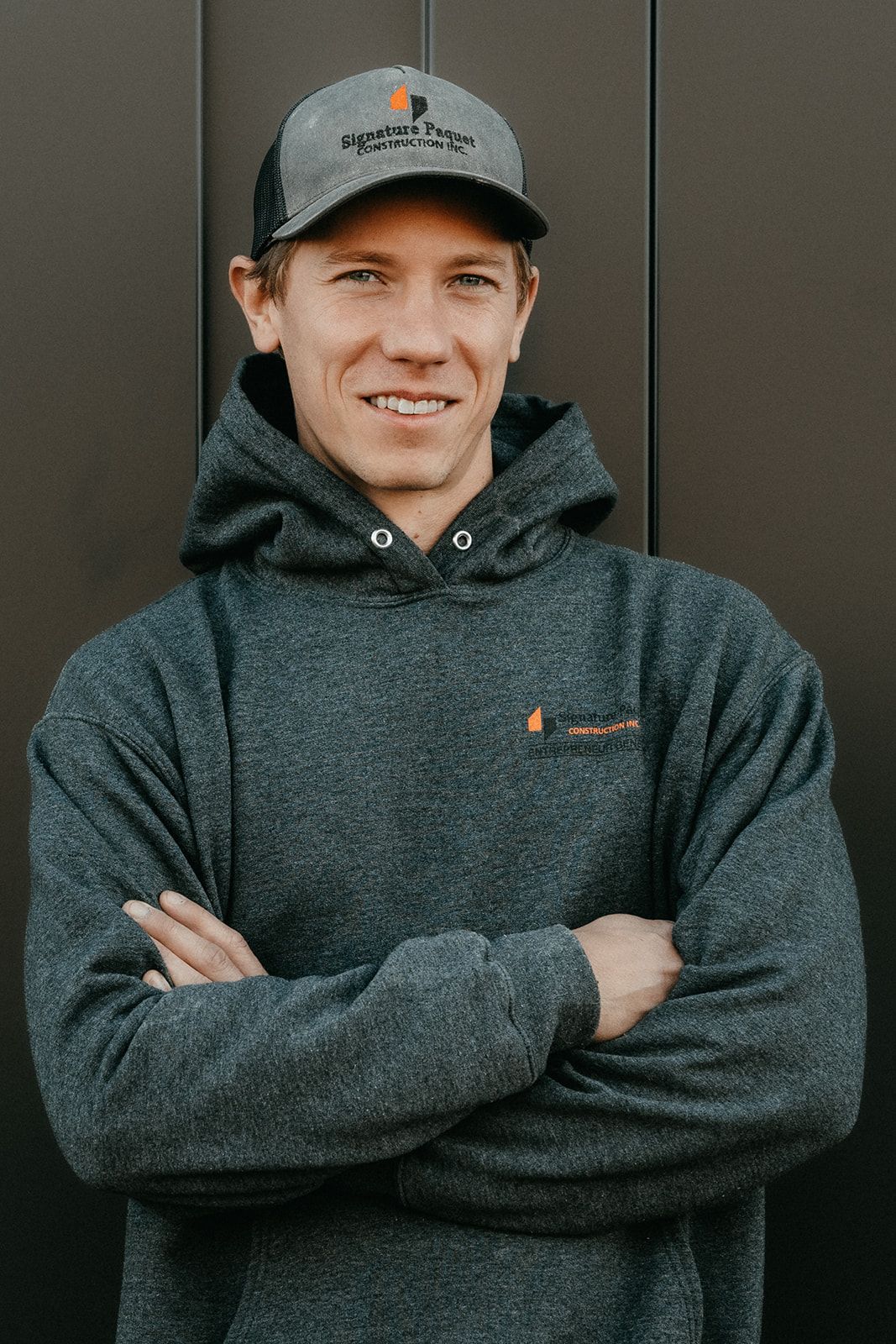 Un homme portant un chapeau et un sweat à capuche est debout, les bras croisés.