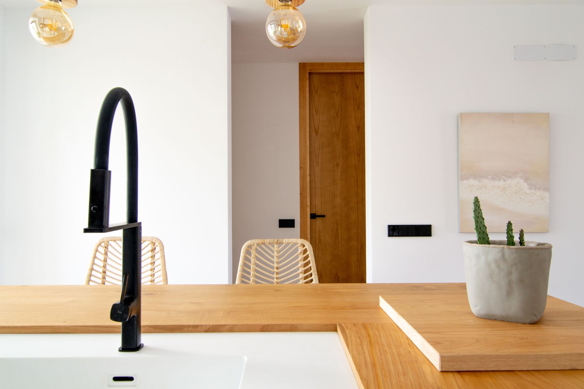 Interior de cocina con grifo negro, encimeras de madera clara, paredes blancas y una suculenta.