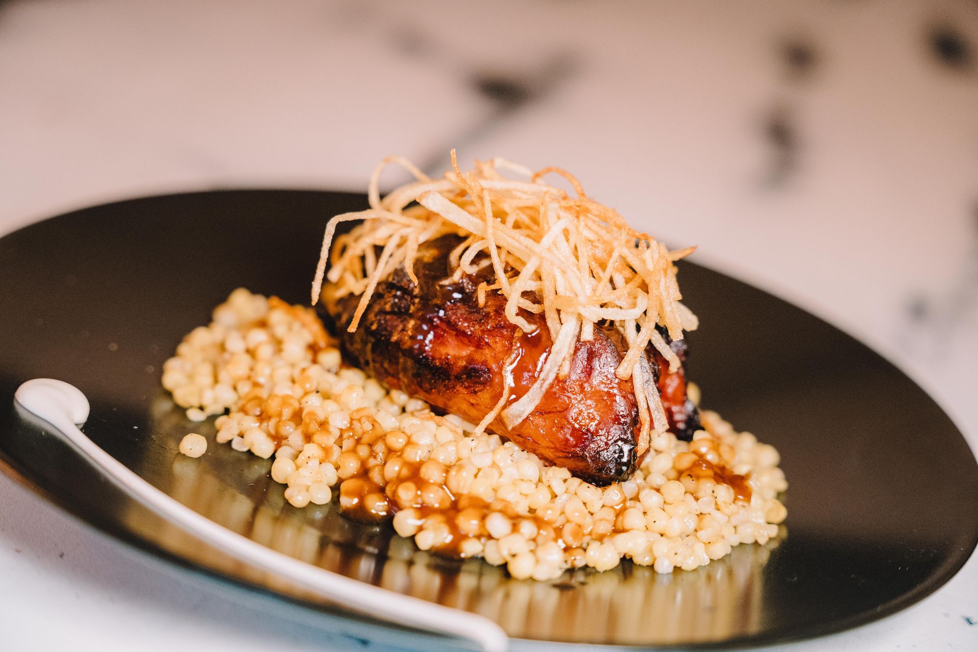 Carne sellada con cubierta crujiente, servida sobre cuscús en un plato negro, fondo de mármol blanco.