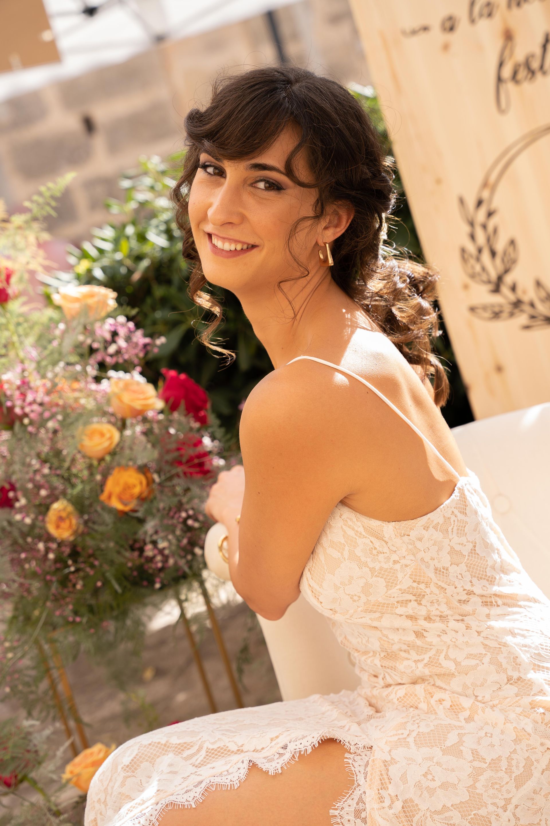 Una mujer con vestido de encaje sonríe al aire libre. Flores y un cartel de madera al fondo.