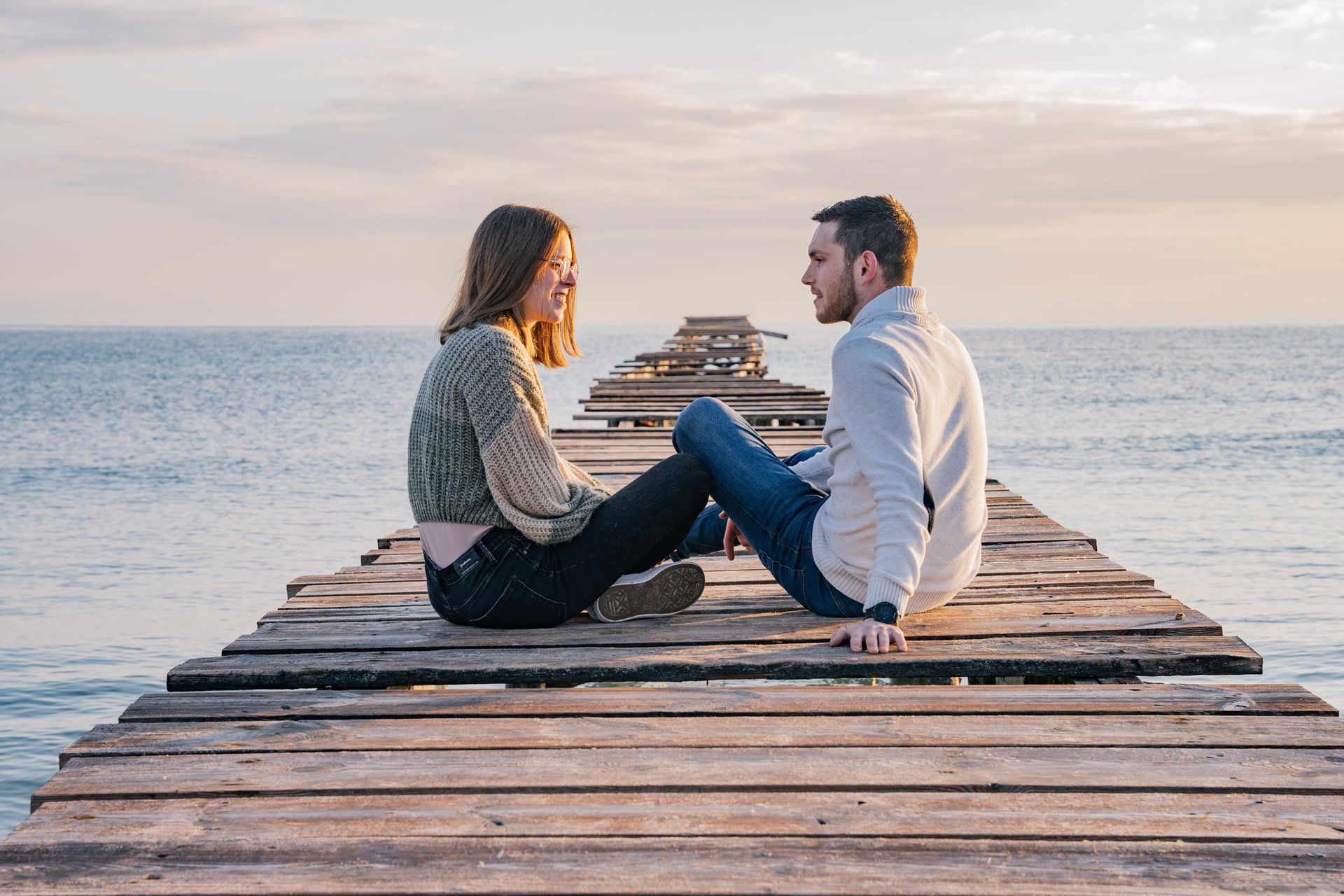 Una pareja se sienta uno frente al otro en un muelle de madera sobre el océano, sonriendo.