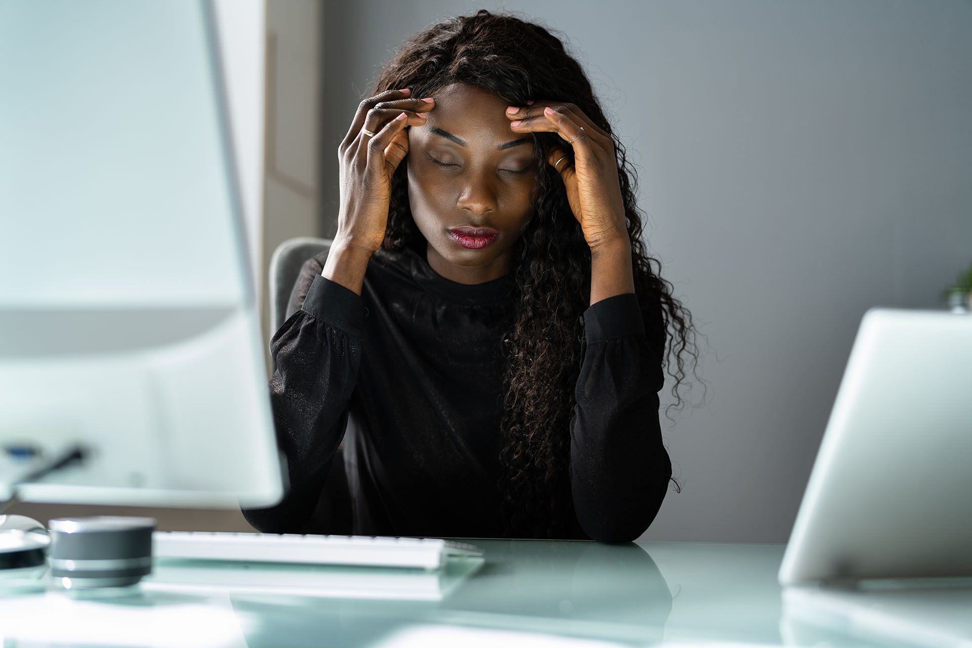 A woman is sitting at a desk with her hands on her head.