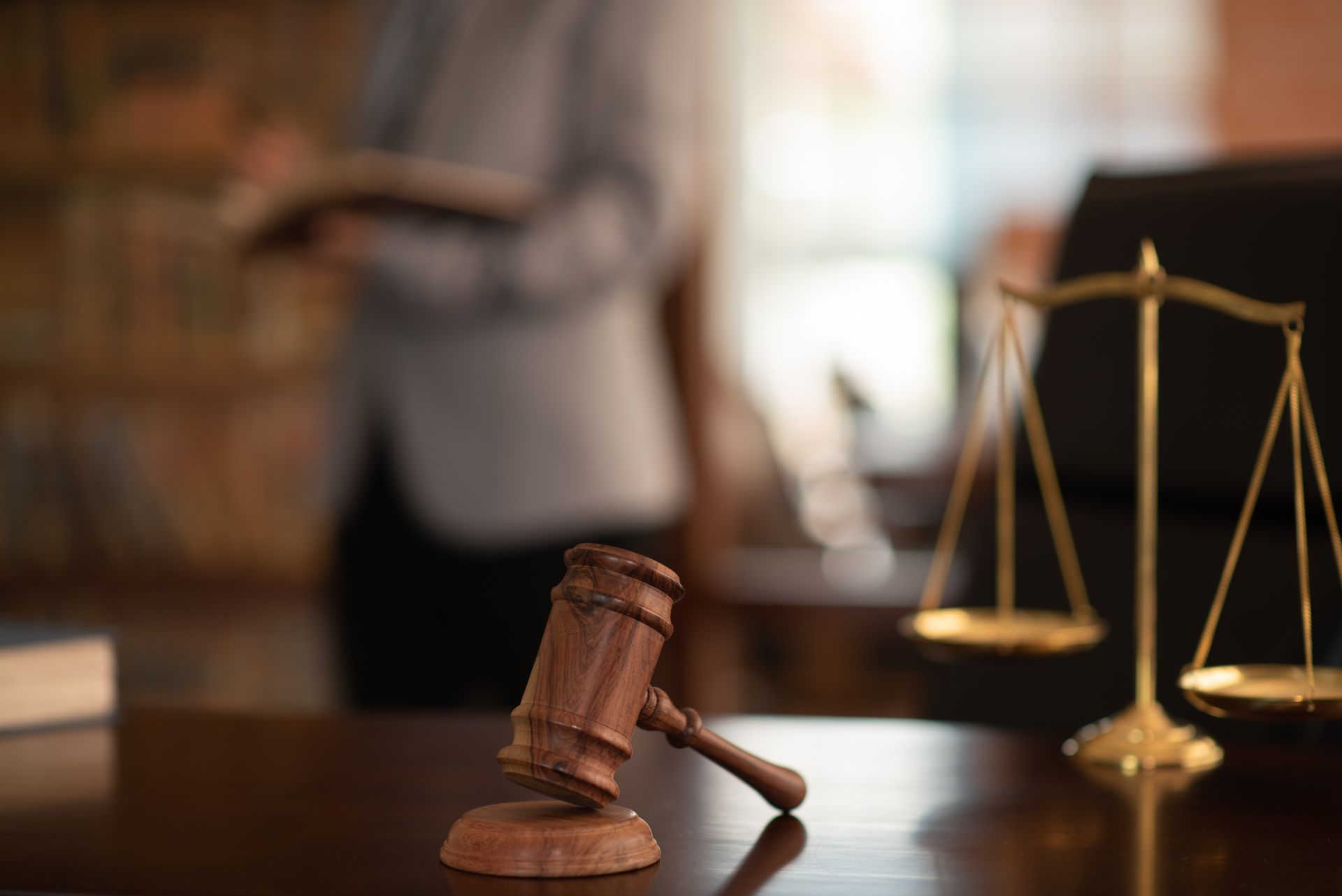 Mazo y balanza de la justicia sobre un escritorio, con una persona sosteniendo un libro en el fondo.