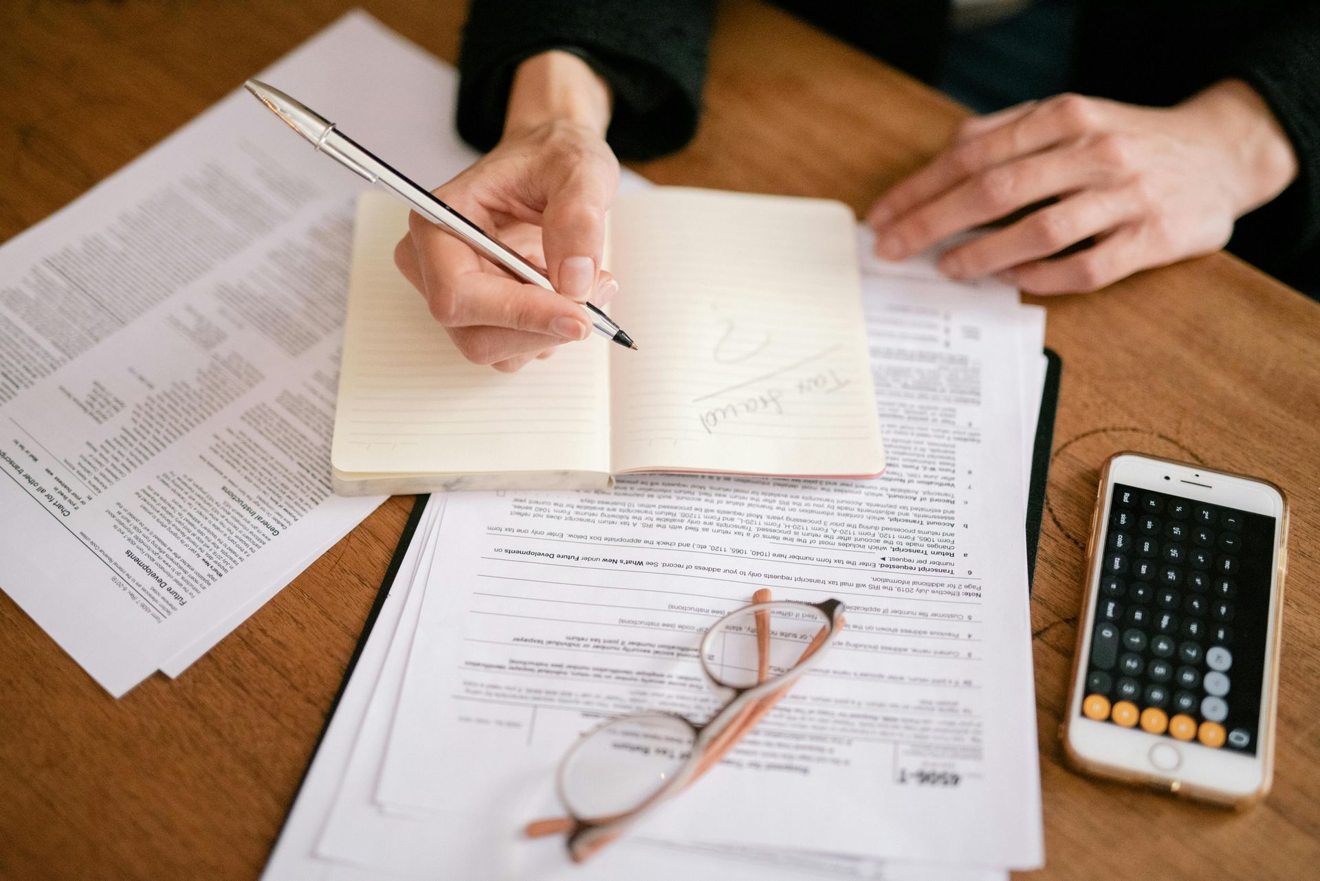 A person writing in a notebook on a wooden desk surrounded by tax forms, glasses, and a smartphone calculator.