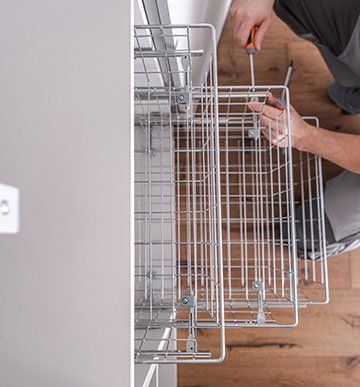 A man is fixing a wire basket with a screwdriver in a kitchen.