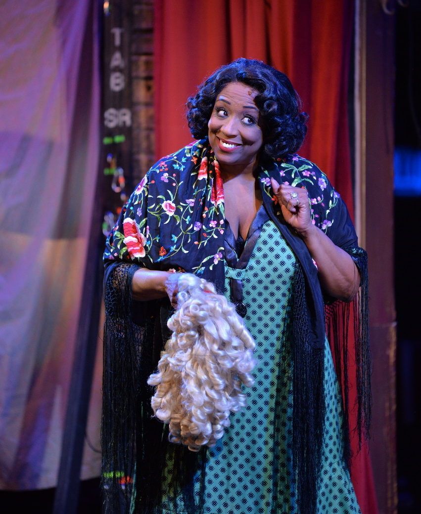 A woman in a patterned dress, shawl, and wig smiles, holding a fluffy blonde accessory.