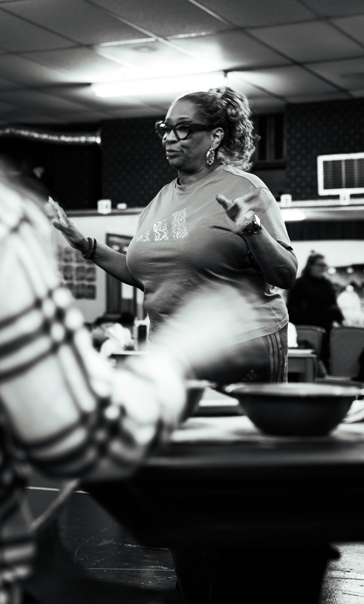 Woman with glasses gestures, speaking. Indoor setting, bowls and person in foreground.