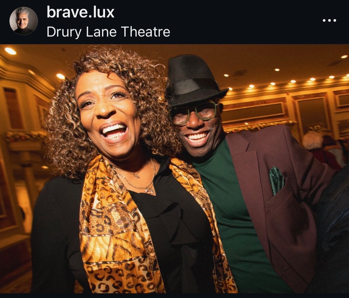 Two people smiling at Drury Lane Theatre. Woman with curly hair and scarf; man in fedora and suit.