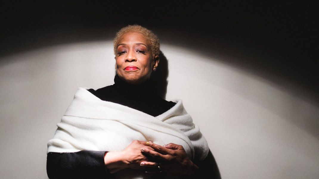 Woman with blonde hair, hands clasped, in white shawl and black turtleneck, set against a shaded wall.