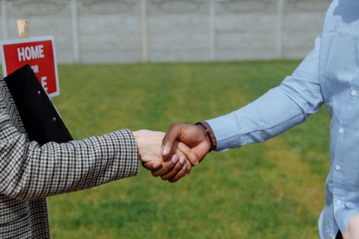 Two people shake hands in front of a house for sale sign on a lawn.