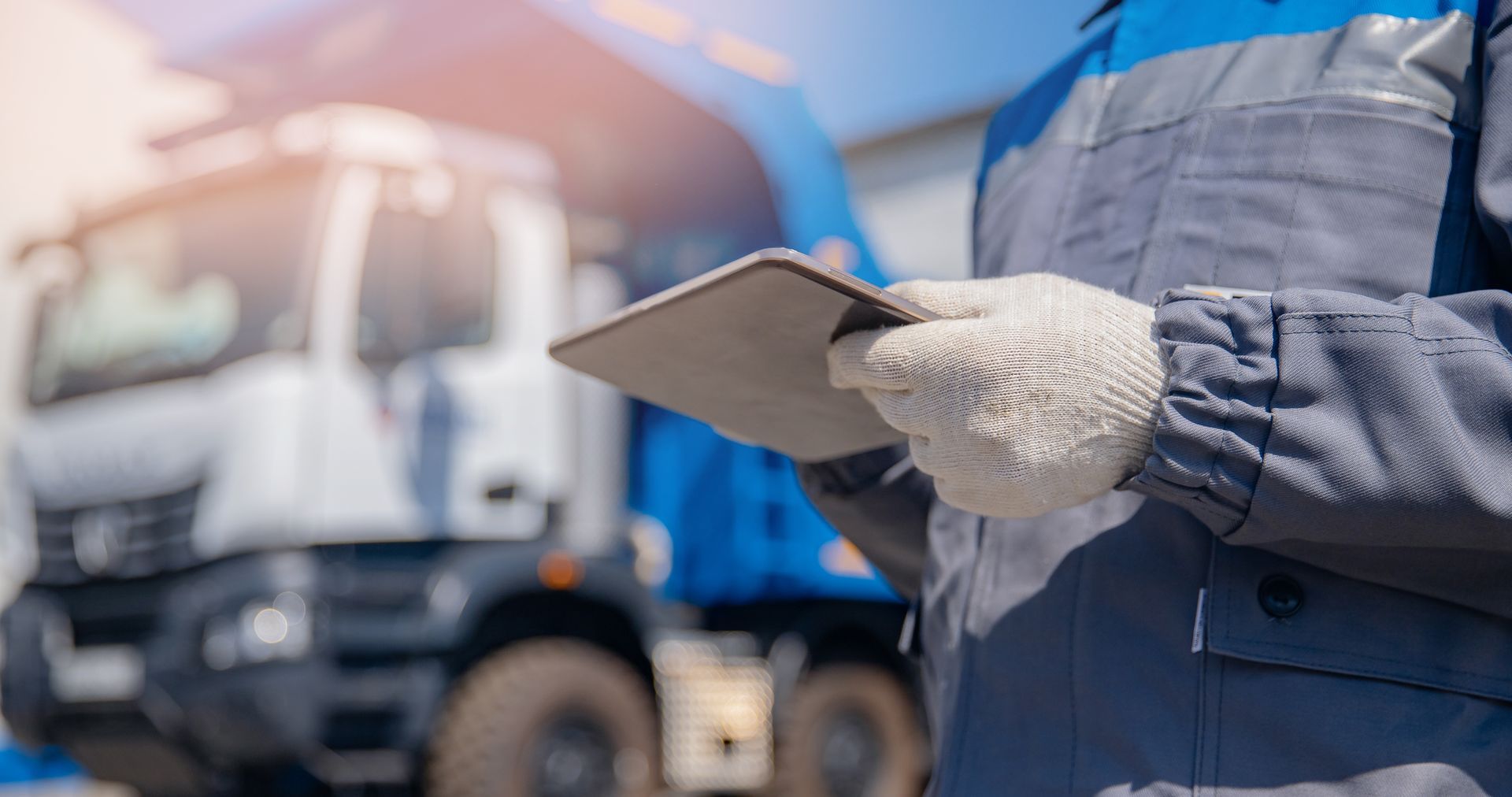 A worker wearing gloves and blue coveralls holds a tablet, standing near a blue and white truck in bright sunlight.