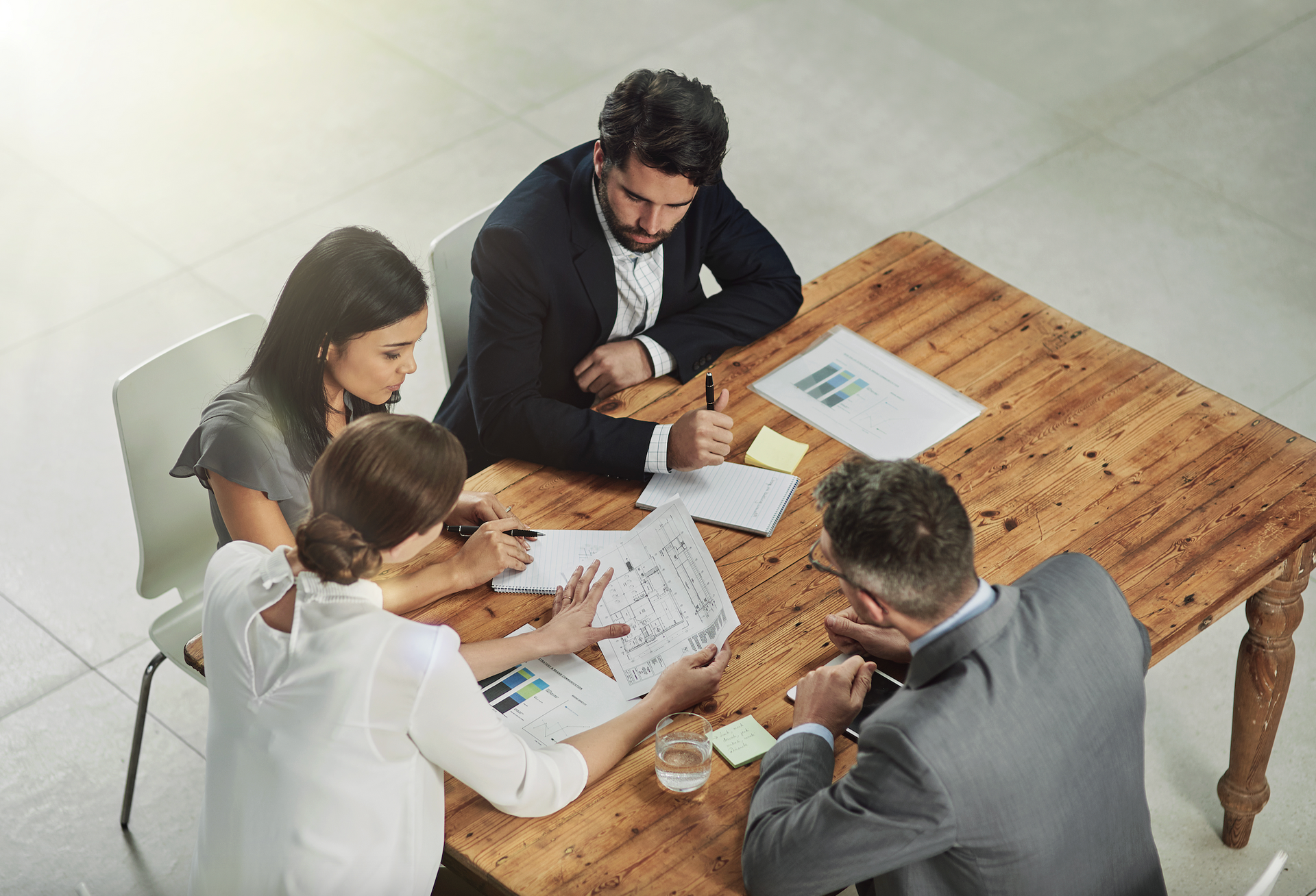 Four business professionals in a meeting around a wooden table, reviewing papers and discussing a project.