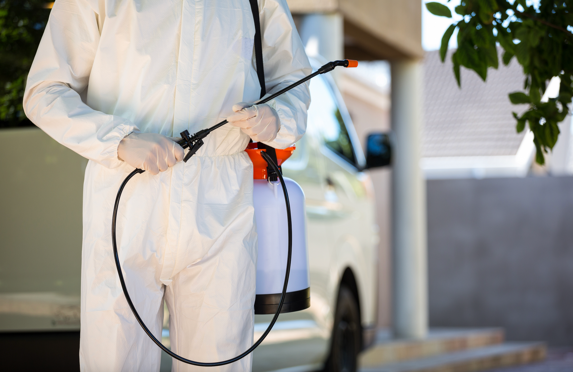 Person in white protective suit holding a sprayer, standing near a vehicle and a house.