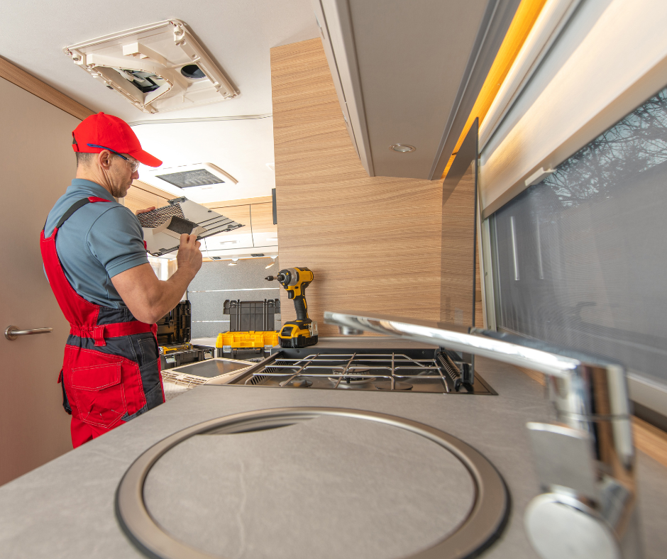 An interior view of an RV showcasing a technician in a red cap, red and gray overalls, working on assembling an RV. The RV kitchenette is equipped with a stainless-steel sink and a stove. Tools like a cordless drill and a toolbox are placed on the countertop, indicating ongoing work. Natural light streams in through the window, with an open roof vent visible above, highlighting a practical and modern RV design.