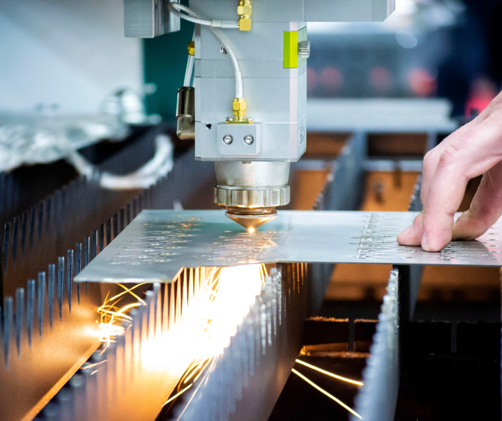 An industrial laser cutter in operation, with a focused beam cutting through a metal sheet, creating bright sparks and a glow from the intense heat. A person's hand is seen close to the cutting area, pointing or adjusting the process, demonstrating the precision work involved. This image highlights the use of specialty gases, which are likely utilized to enhance the laser cutting process, as supplied by a company like Metro Welding Supply Corp. The environment suggests a focus on manufacturing or metalworking where such gases are critical for efficient and high-quality cutting.