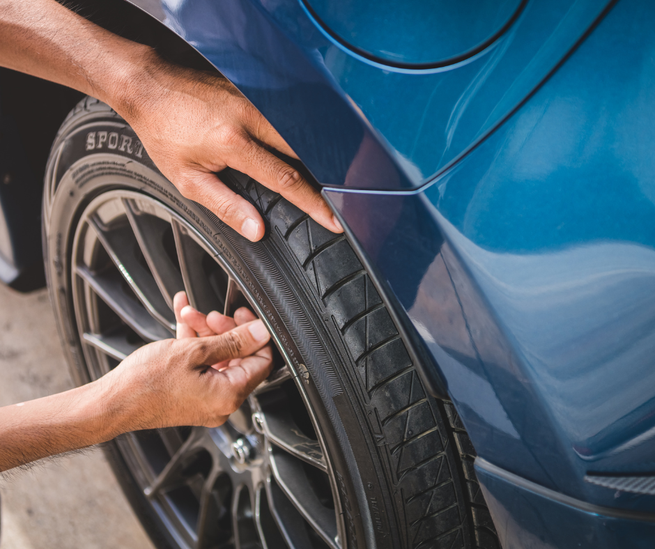 The image shows a person's hands performing a check on the tire of a blue car. The hands are adjusting or inspecting the sidewall of the tire, which is mounted on a sleek, dark multi-spoke wheel. The photo is a close-up, focusing on the interaction between the hands and the tire, with the blue body of the car subtly visible in the background. This indicates a maintenance or safety inspection process, likely being performed by a professional or a diligent vehicle owner.