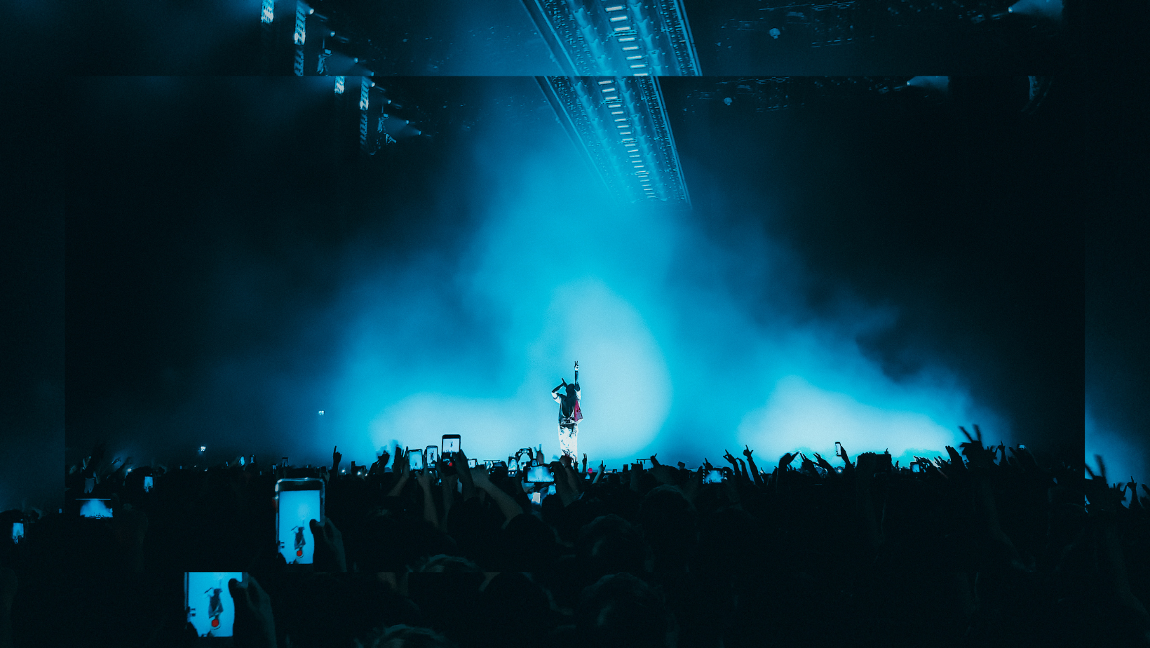 An electrifying concert atmosphere heightened by dense blue fog enveloping the stage, with a dynamic performer engaging the crowd, who are illuminated by stage lights and captured in silhouette. Scores of raised hands and mobile phones recording the moment create a landscape of excitement and shared experience, demonstrating the dramatic impact of specialty gases in entertainment.