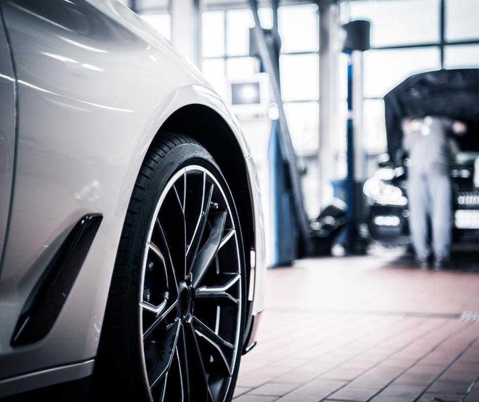 The image displays a close-up of a shiny, white modern car's front wheel and fender, featuring a large, stylish multi-spoke alloy wheel. The car is parked in a bright, clean automotive service center. In the softly focused background, there's another car with its hood open, and a technician is working on it. The environment suggests a high-end dealership or auto shop with large windows allowing natural light to flood the space, highlighting the cleanliness and professionalism of the setting.