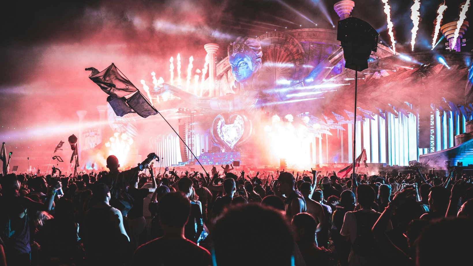 An exhilarating festival scene showcasing a grand stage with a heart-shaped centerpiece, framed by the imposing figure of a deity. The air is alive with a vibrant mix of red and blue lighting, while fireworks and pyrotechnics light up the night sky, creating a visually stunning backdrop. Energetic crowds, some with flags, are bathed in the glow of the spectacle, illustrating the transformative power of specialty gases in creating breathtaking pyrotechnic and atmospheric effects at large-scale events.