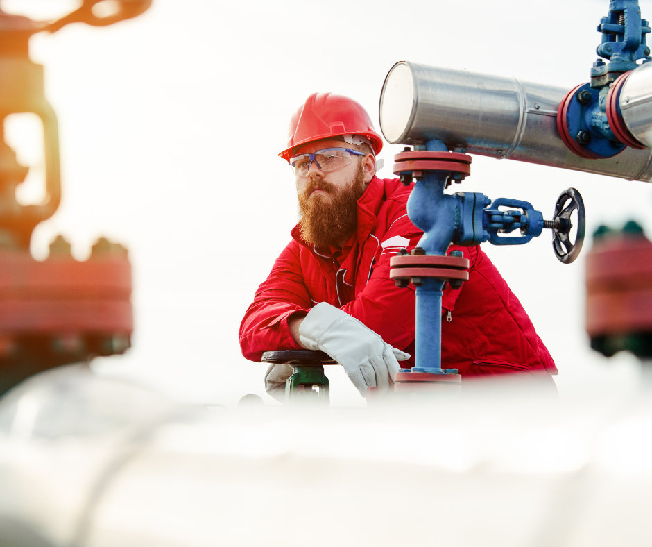 A focused industrial worker in a red uniform and hardhat is monitoring or adjusting a large blue valve on a pipeline. The background is softly blurred, emphasizing the worker in the foreground. He wears protective eyeglasses and a beard, and his attention seems fixed on the equipment, suggesting maintenance, safety inspection, or operational management in an industrial setting.