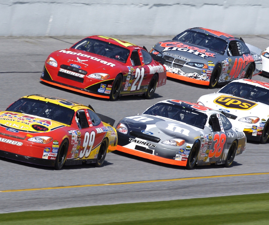 A dynamic and vibrant scene from a stock car race with four brightly colored cars tightly packed and speeding down the track. The lead car, number 99, is vivid yellow with red accents, while the car beside it, number 22, is predominantly red with white and black details. Behind them, car number 40 boasts a fiery orange and black design, and car number 38 is white with yellow and black detailing. All cars are branded with their sponsors' logos and have a model name 'Taurus' visible on their fronts. The background shows a blurred grandstand, indicating high speed, and the focus is on the intensity and competition of the race.