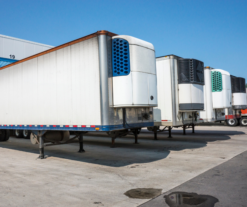 A row of white refrigerated tractor-trailers parked at a loading dock, each equipped with large refrigeration units on their front ends. The units have side vents in varying patterns of blue and teal. The pavement beneath the trailers is marked with occasional stains, and each trailer is hitched to its respective undercarriage with support wheels. The background features the industrial setting of a warehouse with the number 19 visible on one of the doors, indicative of multiple loading bays.