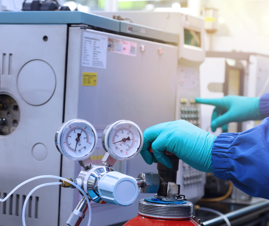A pair of hands wearing blue gloves is adjusting a gas regulator attached to a red gas cylinder. The regulator has two pressure gauges that are used to monitor and control the flow of gas. In the background, there is laboratory equipment, possibly part of a gas chromatography system or other analytical instruments that require a controlled gas supply, such as helium, argon, or nitrogen, which could be provided by a company like Metro Welding Supply Corp. The setting suggests a professional laboratory environment with a focus on precision and safety in handling gases.