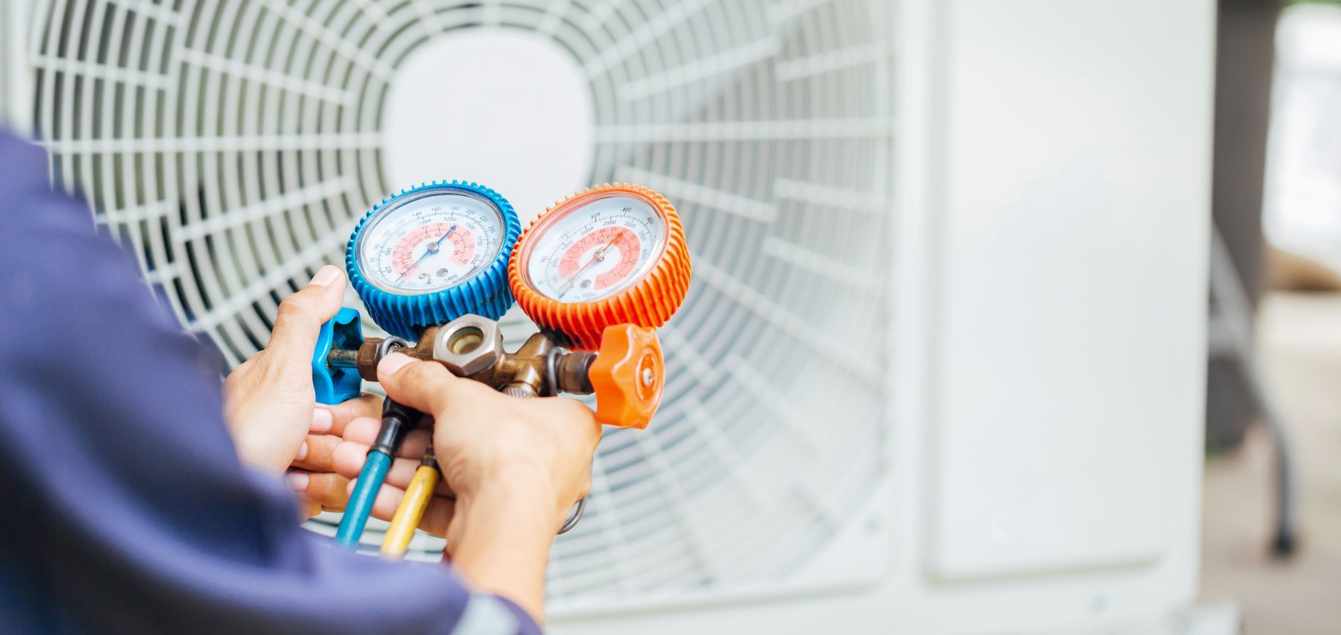 A person servicing an air conditioner uses gauges. The gauges are blue and orange and connected to the machine.