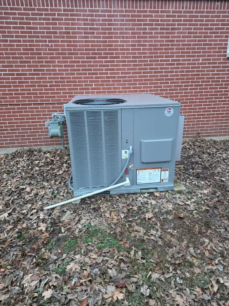A gray HVAC unit with a large fan, next to a brick wall. The unit sits on fallen leaves.