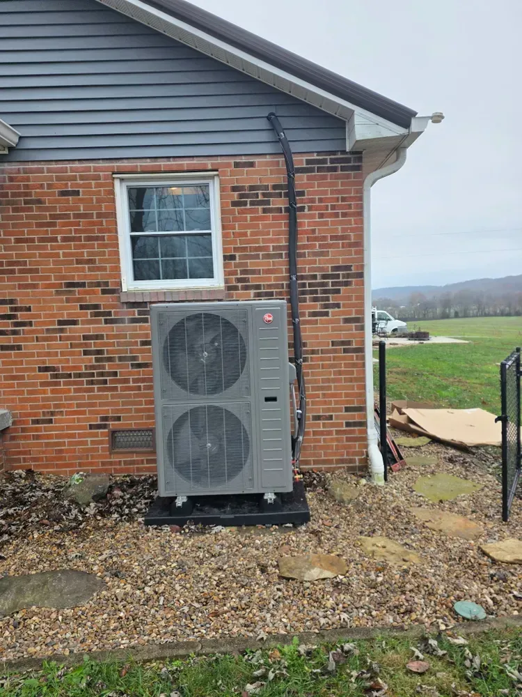 An outdoor heat pump unit next to a brick house. Black pipe runs up the wall.