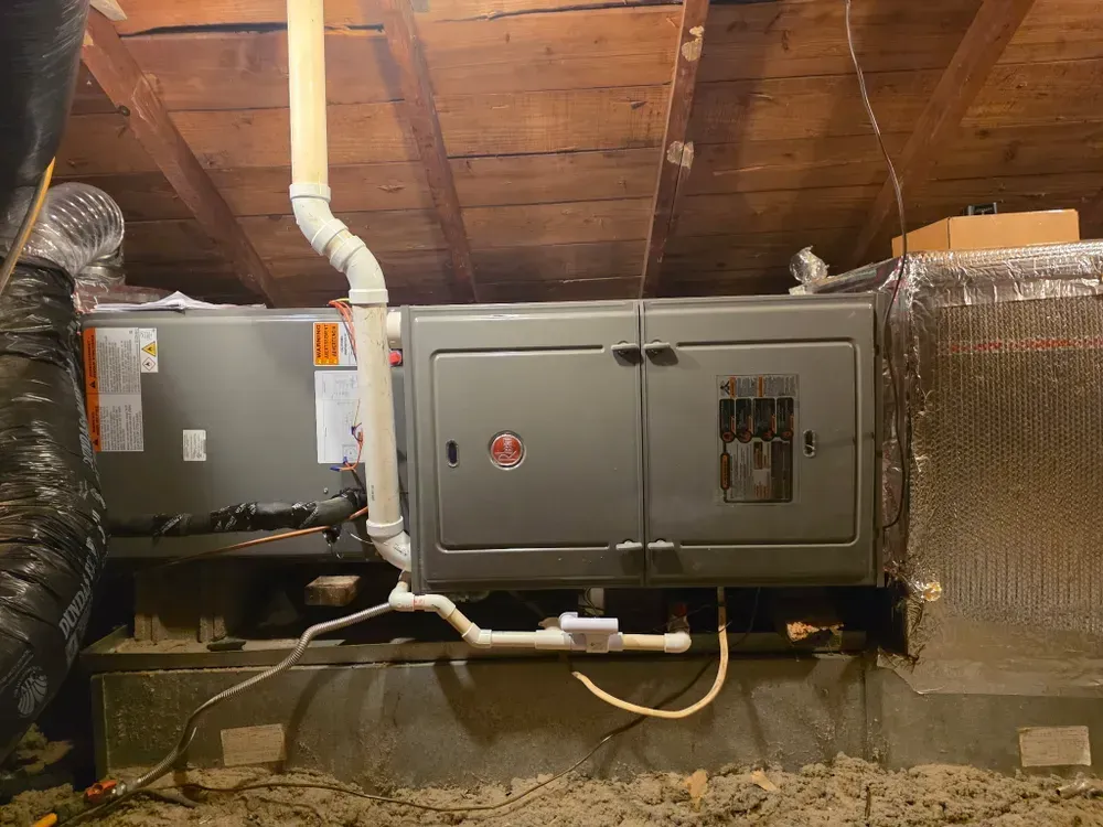HVAC unit in attic with ductwork, electrical panel, and PVC drain pipes. Gray metal unit set on wooden base.