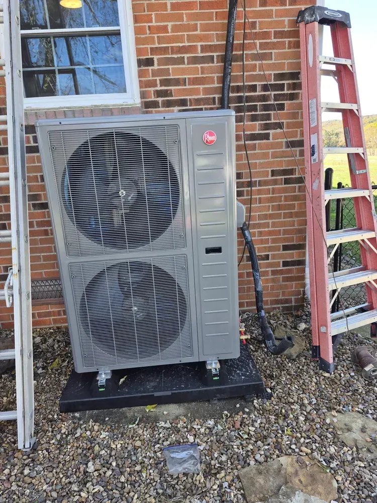 A Rheem heat pump unit installed outside a brick building, with ladders nearby on a gravel surface.
