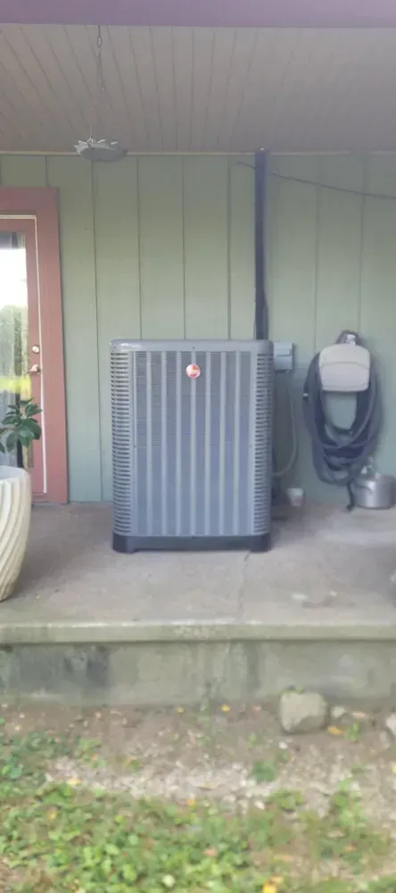 A gray air conditioning unit sits on a concrete step next to a green wall. A hose hangs nearby.