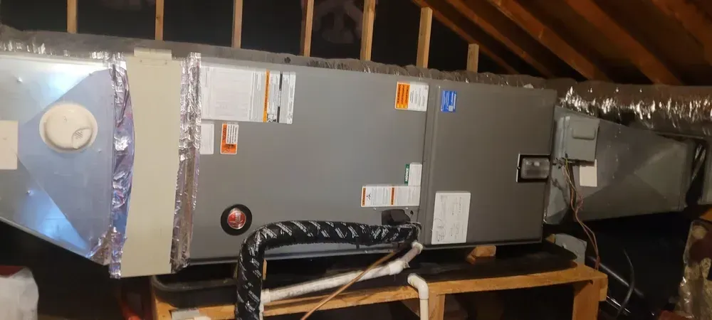 HVAC system in an attic. Ductwork, a furnace, and other components are visible. The system sits on a wooden platform.