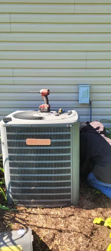 A person working on an outdoor air conditioning unit next to a house with tan siding. A drill sits on top.