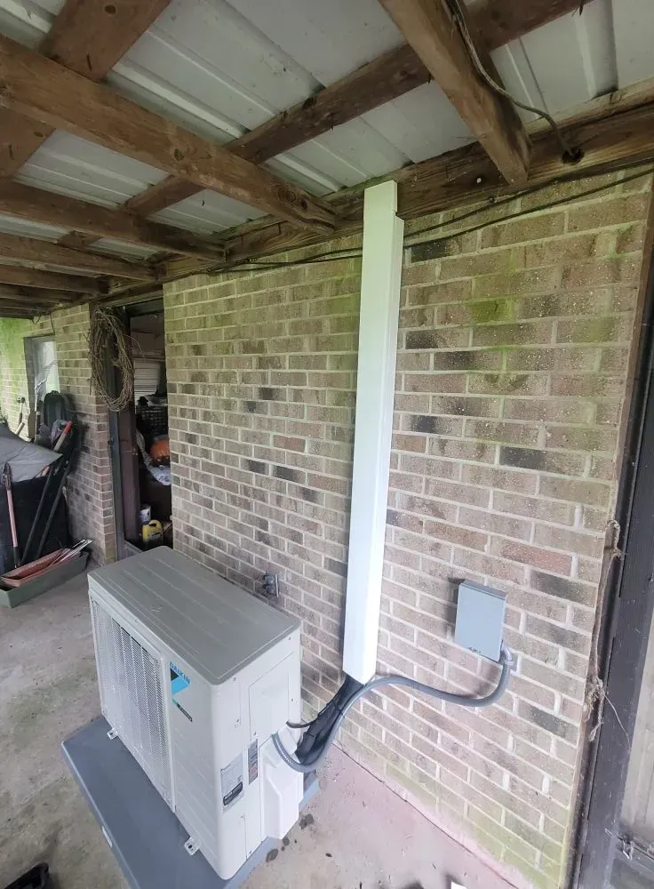 An air conditioning unit on a platform next to a brick wall with a conduit running up to the ceiling.