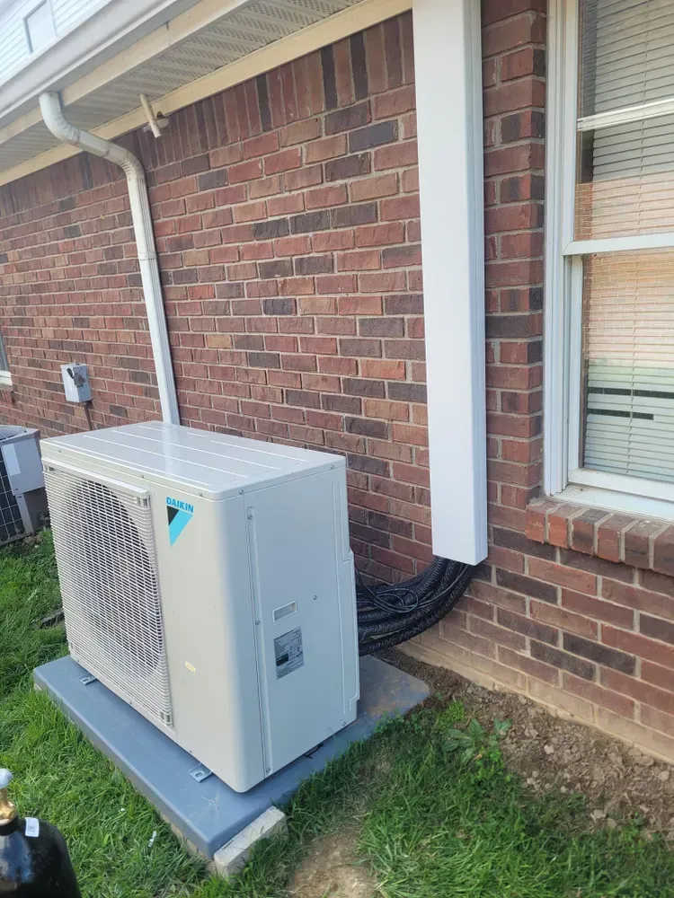Air conditioning unit outside a brick building, with a white conduit running up the wall.