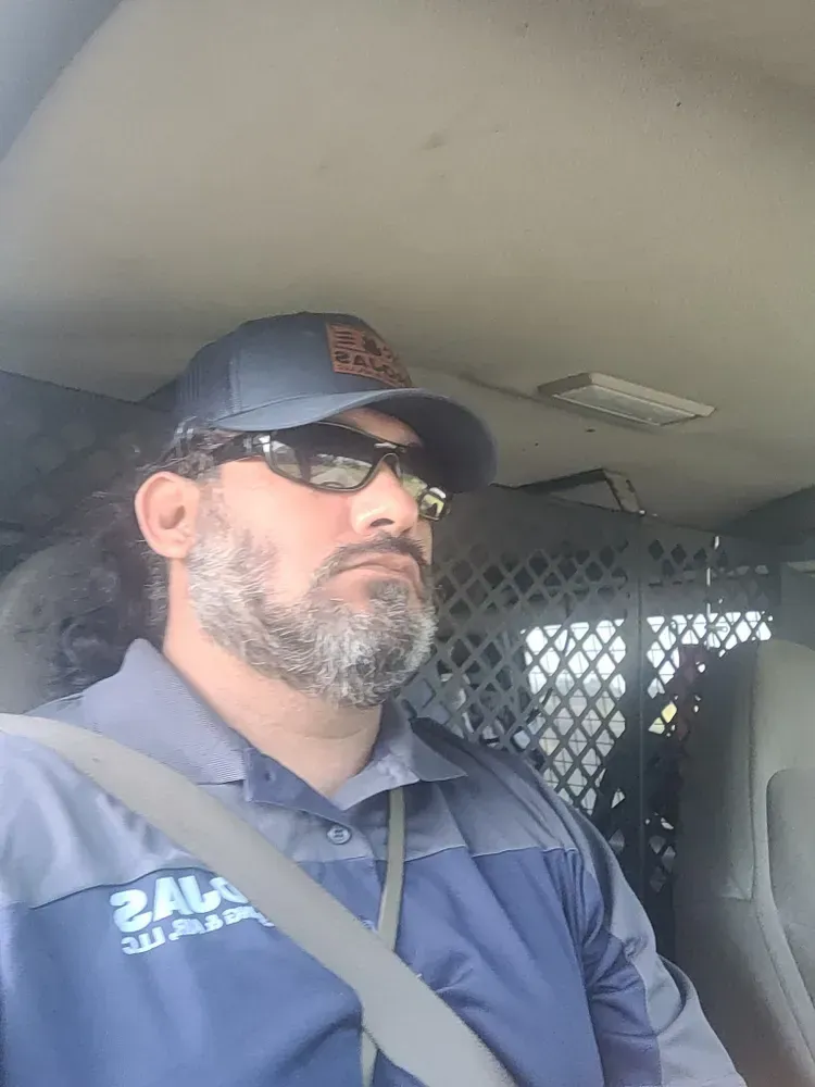Man in cap and sunglasses in a vehicle, wearing a blue work shirt, looking to the side.