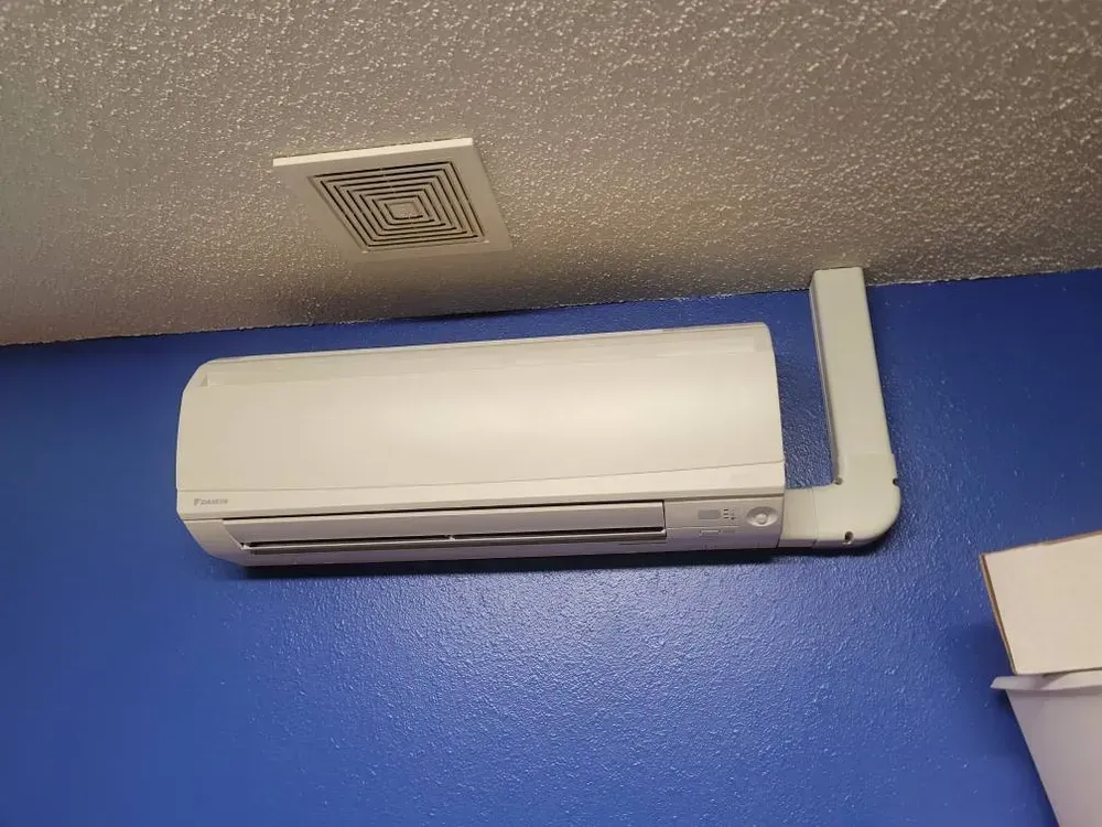 White air conditioner unit mounted on a blue wall, with a ceiling vent above.