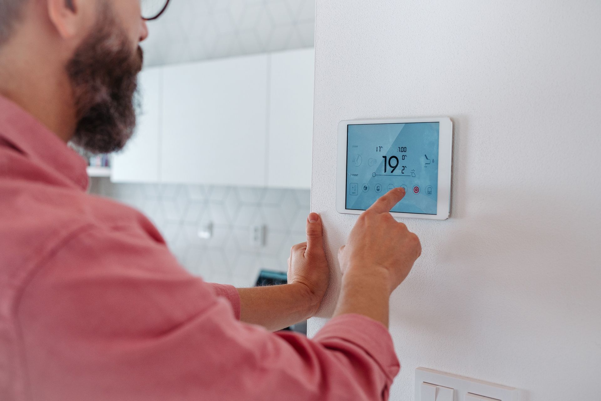Man in pink shirt adjusting a digital thermostat on a white wall in a modern kitchen.