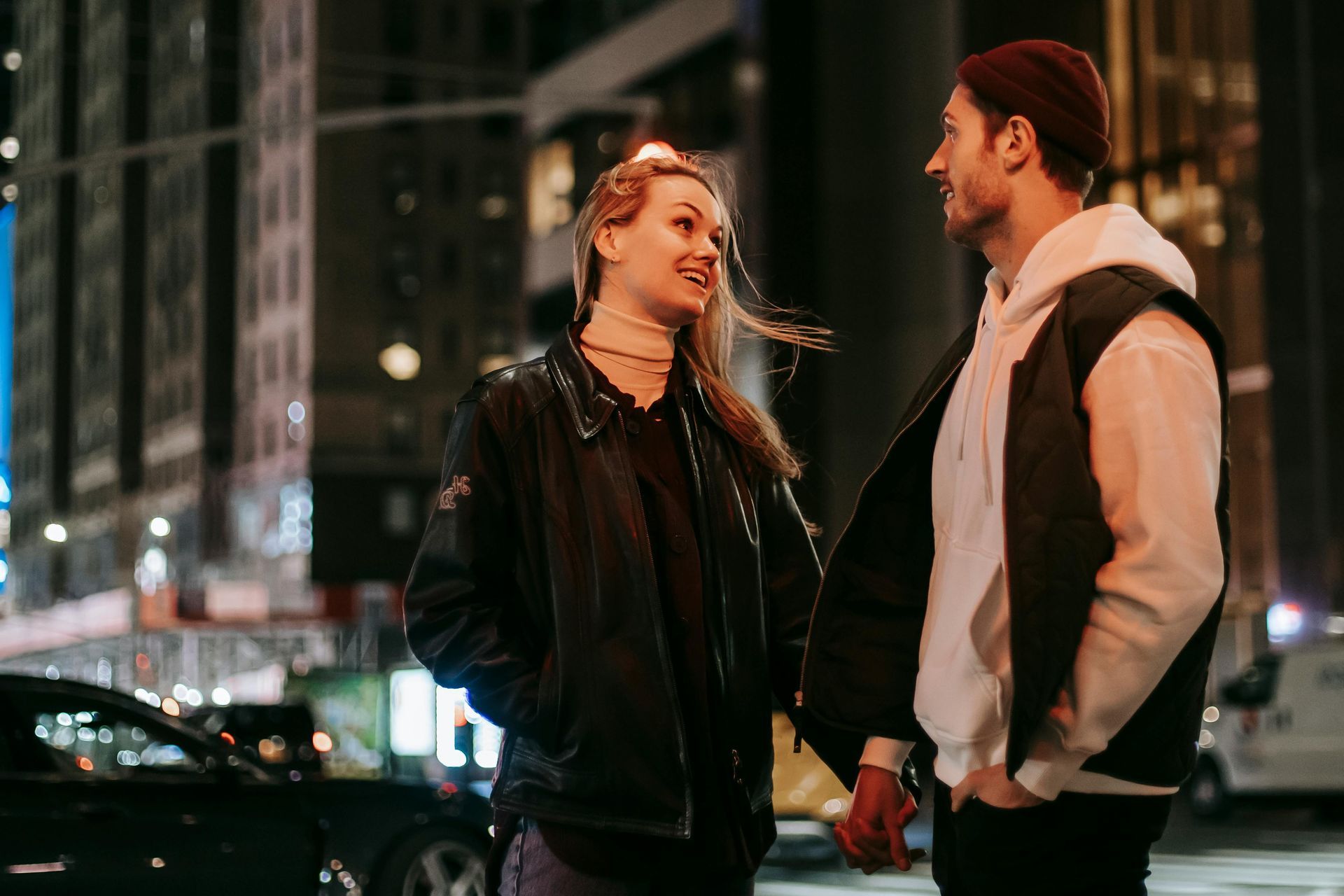 A man and a woman are holding hands and looking at each other on a city street at night.