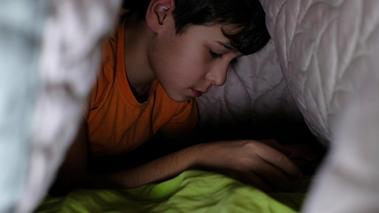 A person in an orange shirt lying under a blanket, looking down at a hand held in the dark.