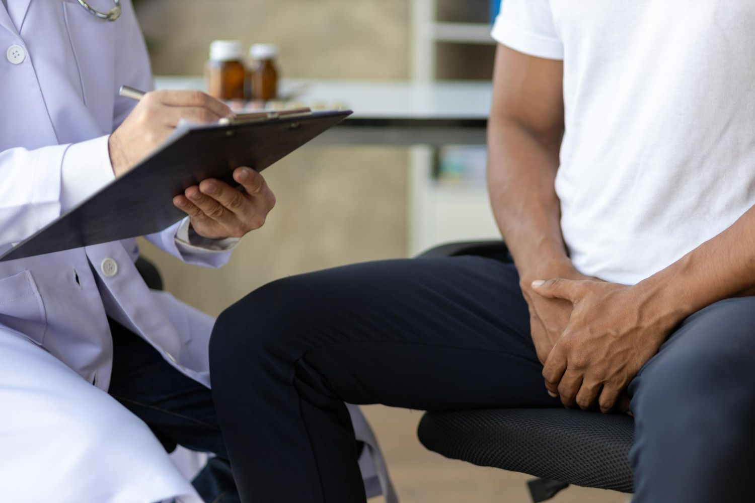 A doctor in a white coat writes on a clipboard while a patient sits nearby holding their hands over their groin area.